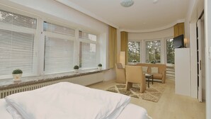 Et hotelværelse med seng, skrivebord, stol, tv og store vinduer med persienner.