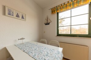 A room with a table, chairs, a window with curtains, a radiator, and wall decorations including framed pictures and a model ship.