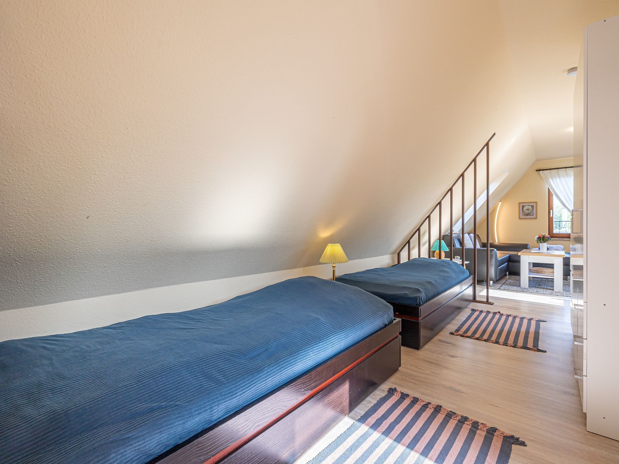 Ein kleiner, kompakter Raum mit zwei Einzelbetten, Holzboden, einer Treppe zu einer oberen Ebene und einem kleinen Tisch mit Lampe.