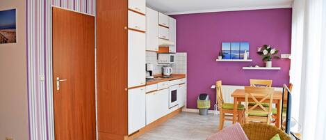 A compact living space with a kitchenette, a dining area, and a seating area with a wicker chair and cushions.