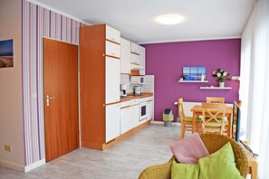 Un espace de vie compact comprenant un coin cuisine, un coin repas et un coin salon avec un fauteuil en osier et des coussins.