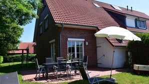 Ett hus med ett rött tegeltak, en fasad av tegel, en täckt uteplats med ett bord och stolar samt ett vitt parasoll.
