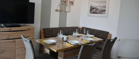 A dining area with a wooden table set for four, a television on a wooden cabinet, and framed pictures on the wall.