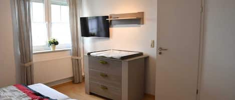 Un dormitorio con una cama, un tocador, un televisor montado en la pared, una ventana con cortinas y una puerta.