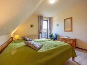 A bedroom with a bed, a chair, a nightstand, a window with curtains, and a radiator.