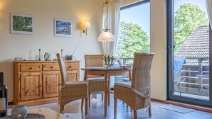 Een eethoek met rieten stoelen, een houten tafel en een kast. Er is een raam met uitzicht op bomen en een gebouw.