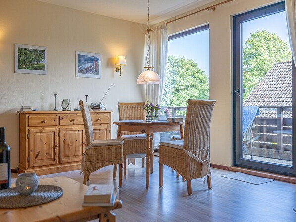 A dining area with wicker chairs, a wooden table, and a cabinet. There is a window with a view of trees and a building outside.