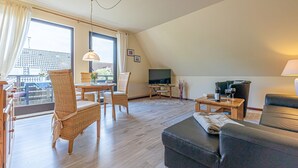Een ruime woonkamer met een groot raam, een televisie, een bank en een eethoek met rieten stoelen.