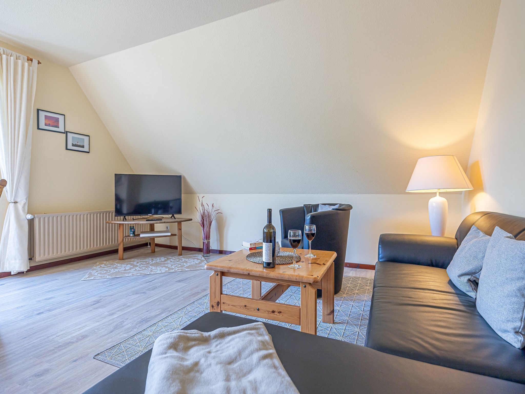 Un salon chaleureux avec un téléviseur, une table basse en bois sur laquelle se trouvent une bouteille et des verres, un canapé en cuir noir et une lampe.