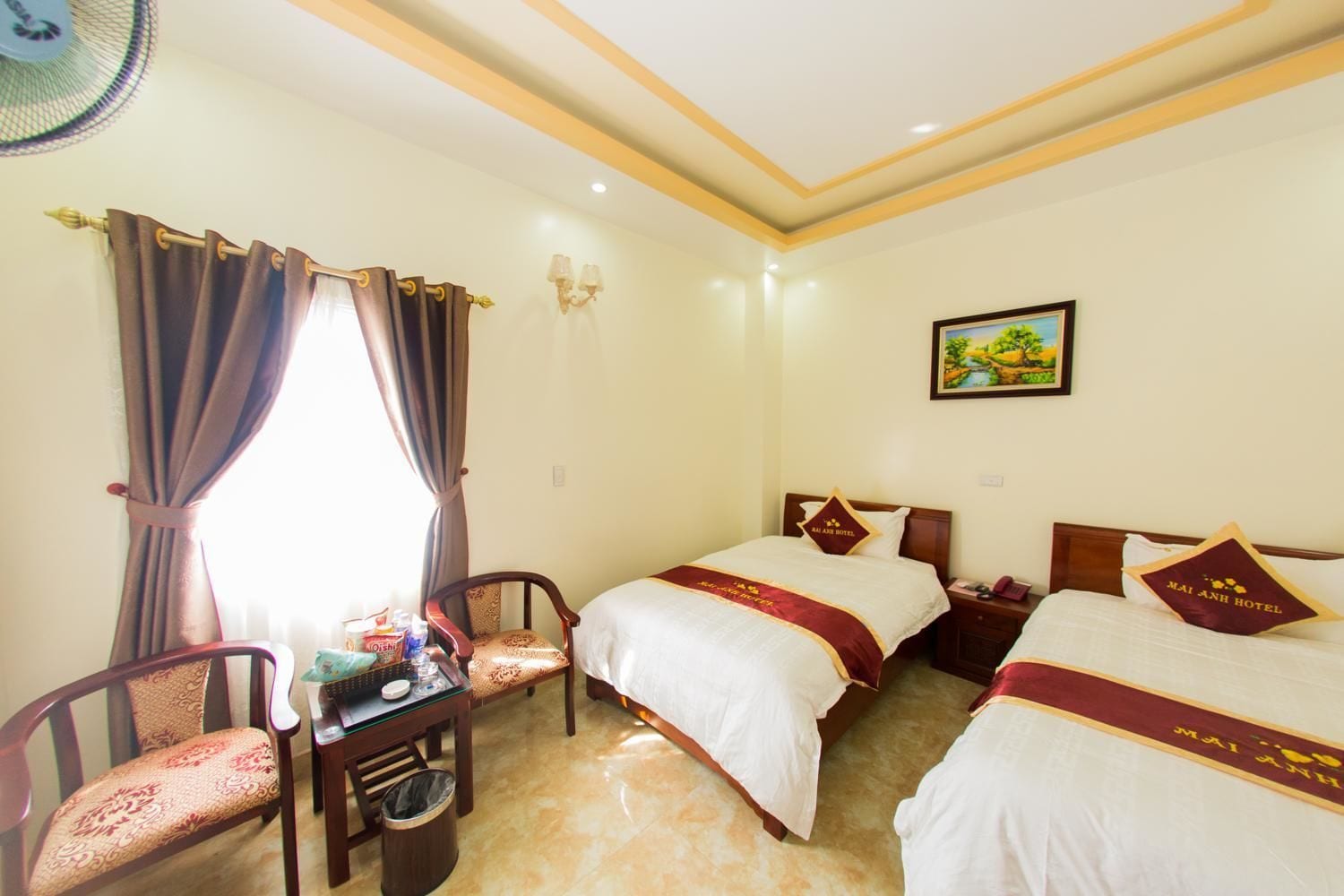A hotel room with two beds, a chair, a small table, and a wall-mounted fan.