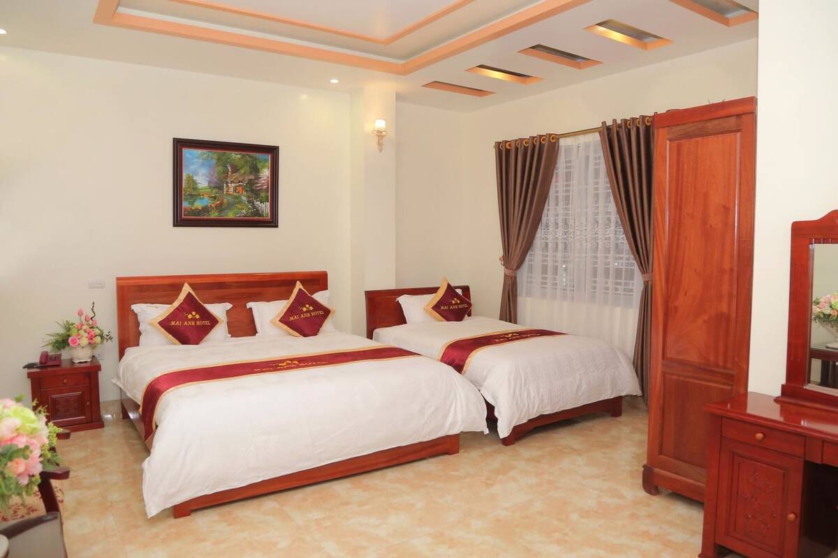 A hotel room with two beds, a wooden headboard, a nightstand with a lamp, a painting on the wall, and a window with curtains.