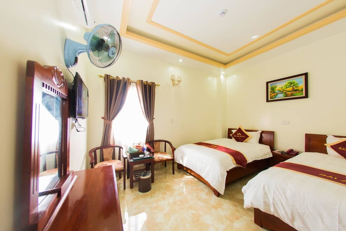 A hotel room with two beds, a desk, a chair, a fan, and a painting on the wall.
