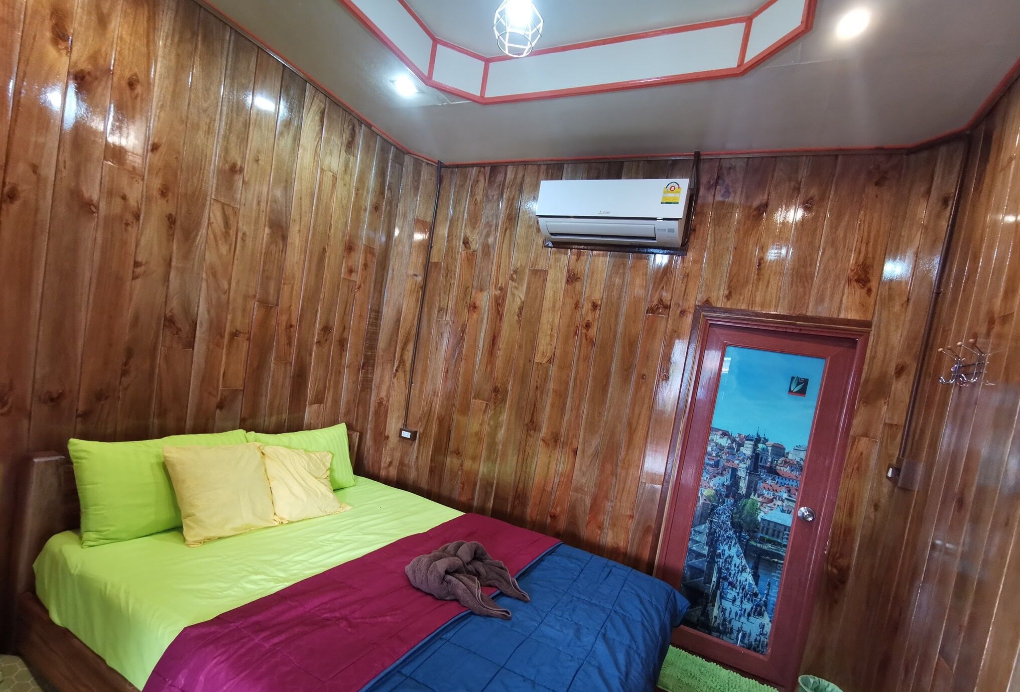 A bedroom with wooden paneling, a bed with green and yellow bedding, a brown blanket, and a window with a cityscape view.