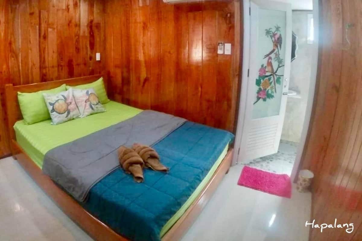 A wooden-paneled room with a bed, green and blue bedding, and an air conditioning unit.