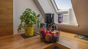 Une cuisine avec un plan de travail en bois, un pot de basilic, une machine à café, un panier de fruits et une fenêtre donnant sur le ciel.
