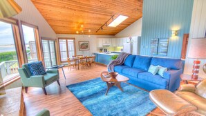 A spacious living room with a blue sofa, green armchairs, and a coffee table. There is a dining area with a table and chairs, and a kitchen in the background.