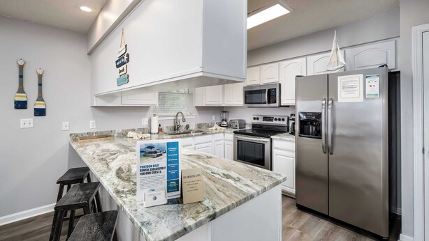 A modern kitchen with white cabinets, a stainless steel refrigerator, a microwave, and a granite countertop. There are two wooden stools and a sign on the counter.
