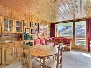 A wooden cabin interior with a dining table, chairs, a sofa, and a kitchen area with cabinets and a window overlooking a mountainous landscape.