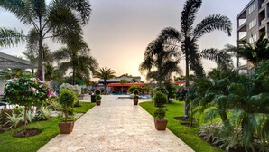 Un chemin pavé mène à un bâtiment doté d’un toit rouge, entouré de palmiers et de diverses plantes.