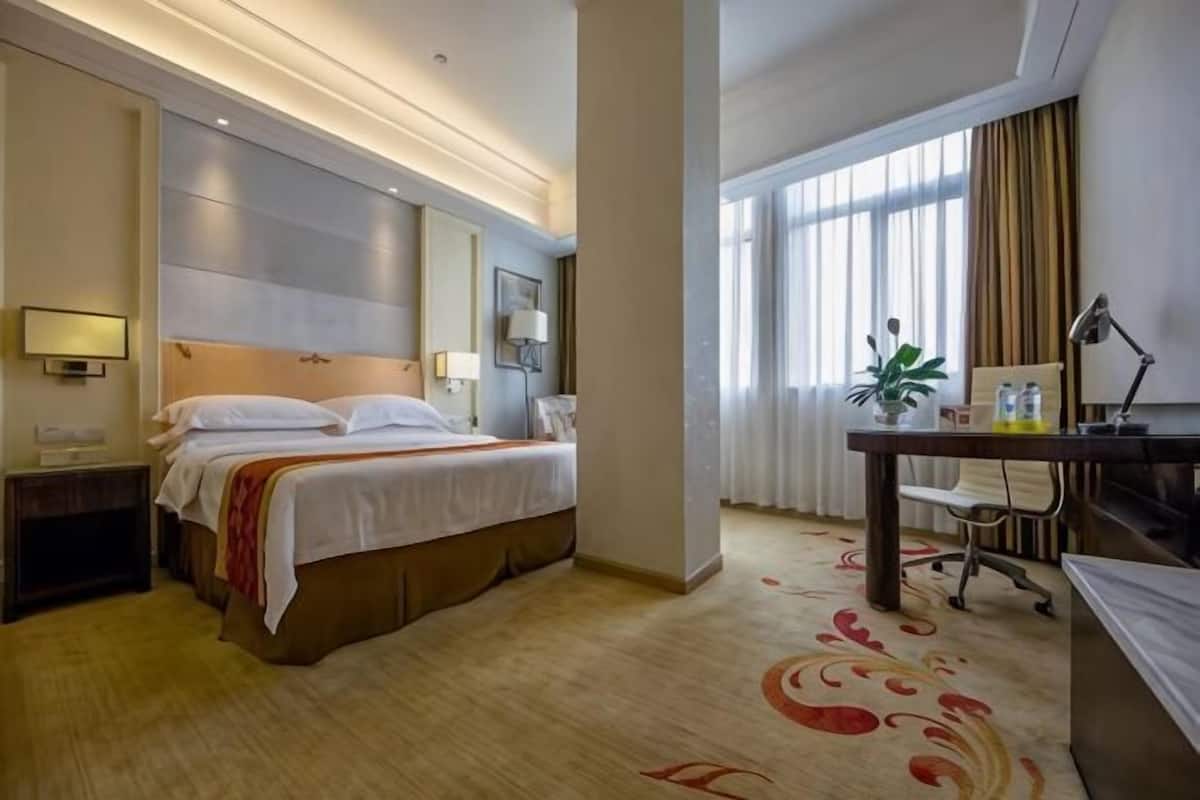 A hotel room with a bed, desk, chair, lamp, and large windows.