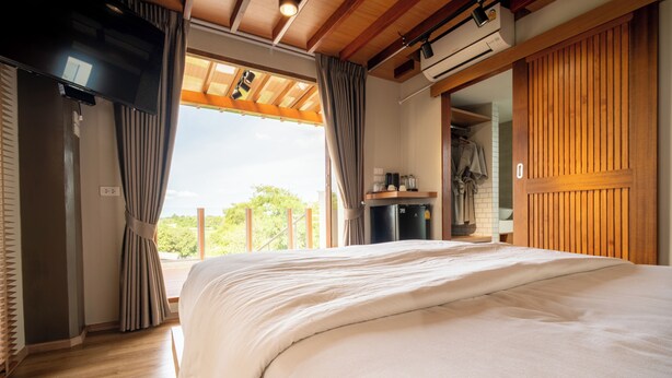 A bedroom with a large bed, wooden ceiling beams, a built-in air conditioning unit, a small kitchenette, and a view through a window with curtains.