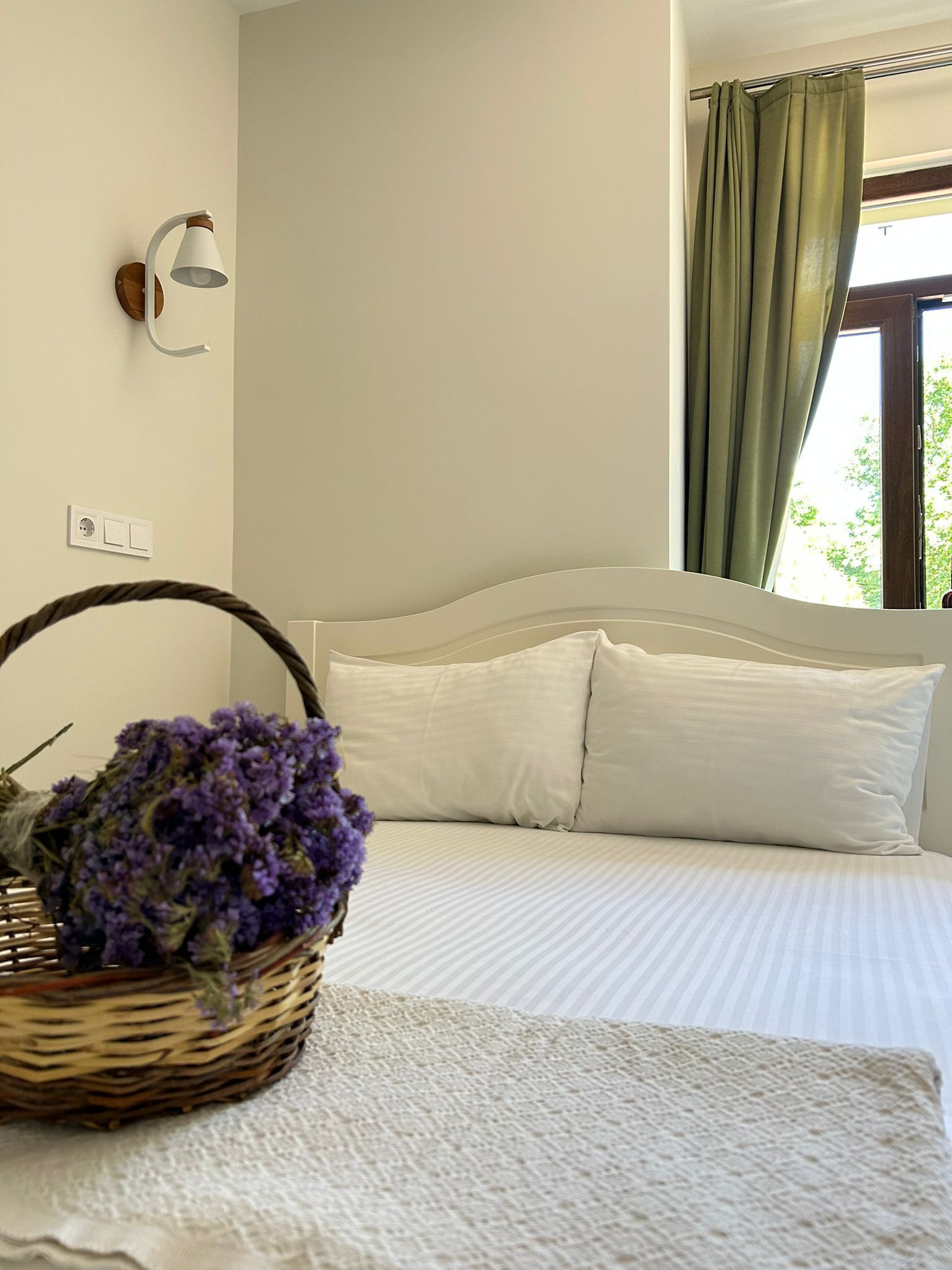 A bedroom with a bed, white pillows, a white blanket, a wall-mounted lamp, a window with green curtains, and a wicker basket with purple flowers.