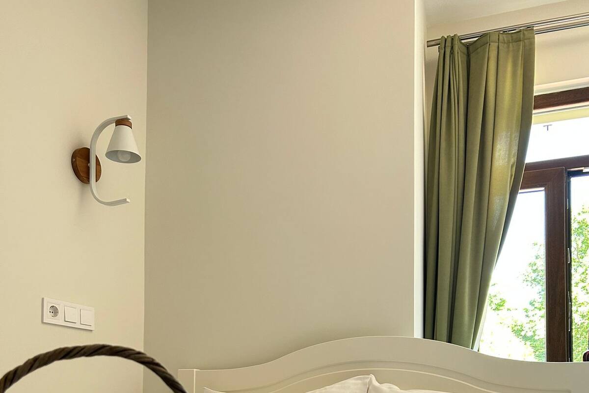 Un dormitorio con cama, almohadas blancas, manta blanca, lámpara de pared, ventana con cortinas verdes y una cesta de mimbre con flores púrpuras.