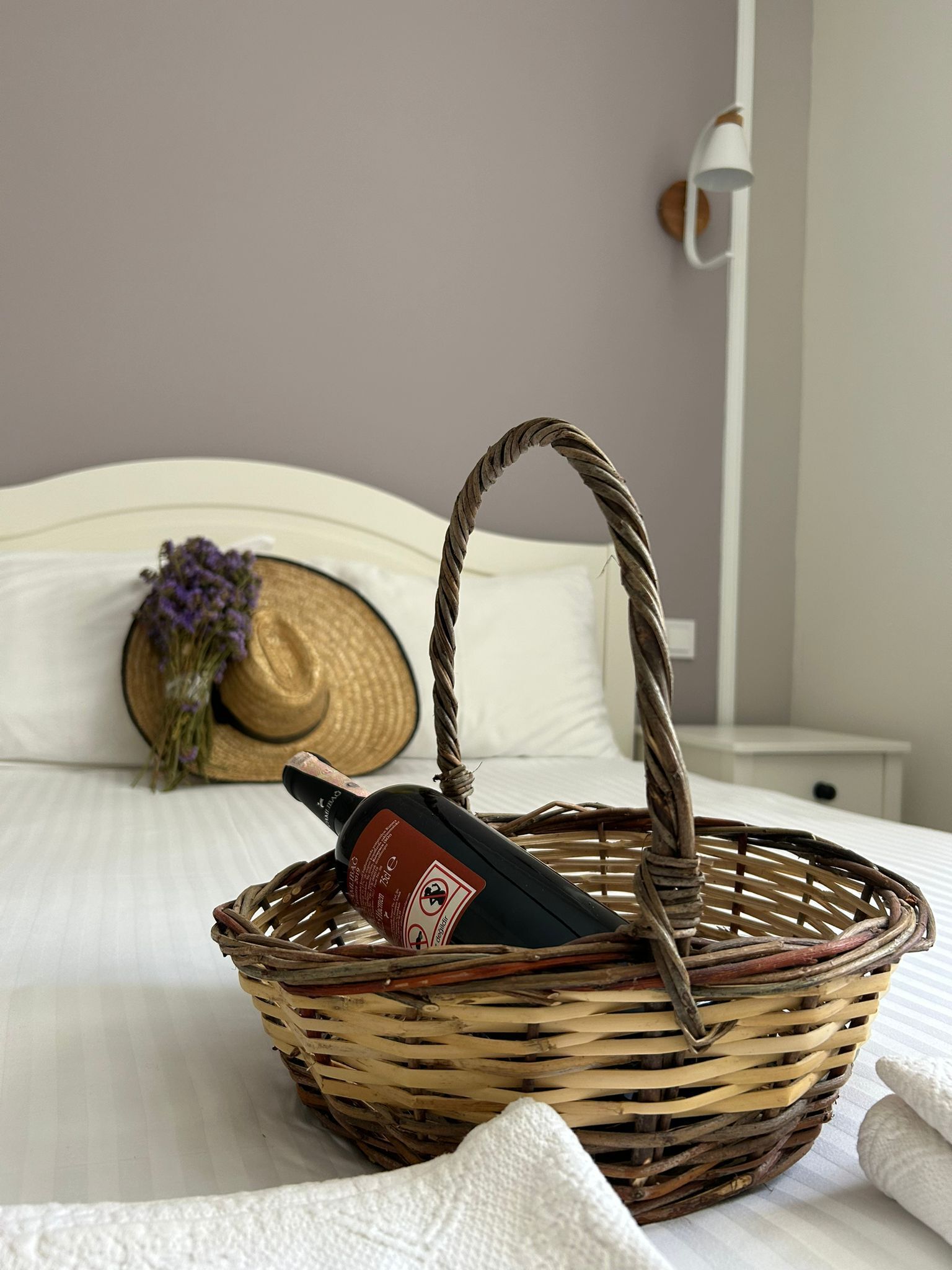 A wicker basket with a bottle on a bed, a straw hat, and a wall-mounted lamp.