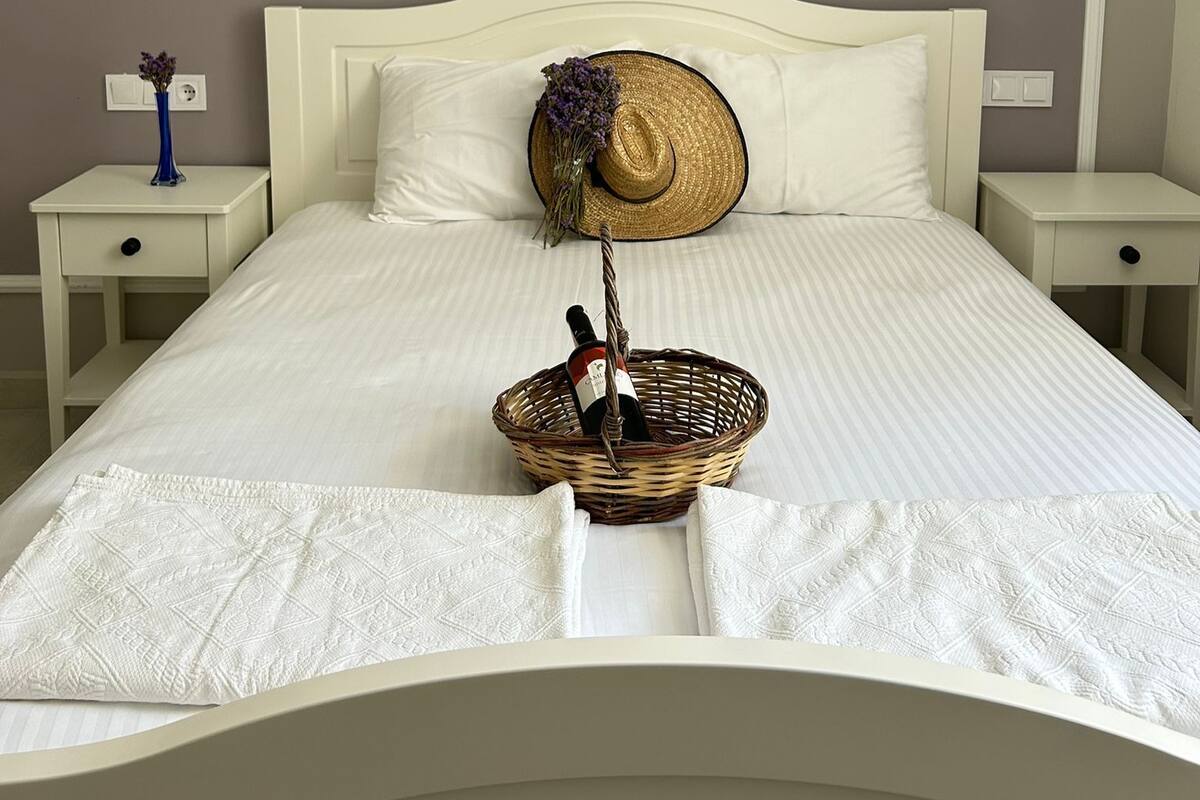 Una cama bien hecha con cabecera blanca, una cesta tejida con una botella y un sombrero de paja sobre la almohada. Hay dos mesitas de noche con lámparas y un jarrón de flores en una de ellas.