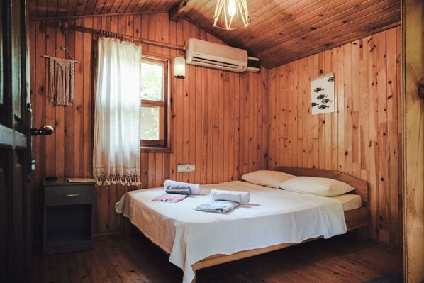 A wooden room with a bed, a window, an air conditioner, and a wall-mounted light fixture. - Base Bungalows (Kumluca)