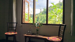 Una habitación con un ventanal grande, dos sillas con asientos de tejido, una mesita con un florero de cristal y vistas a un follaje exuberante.