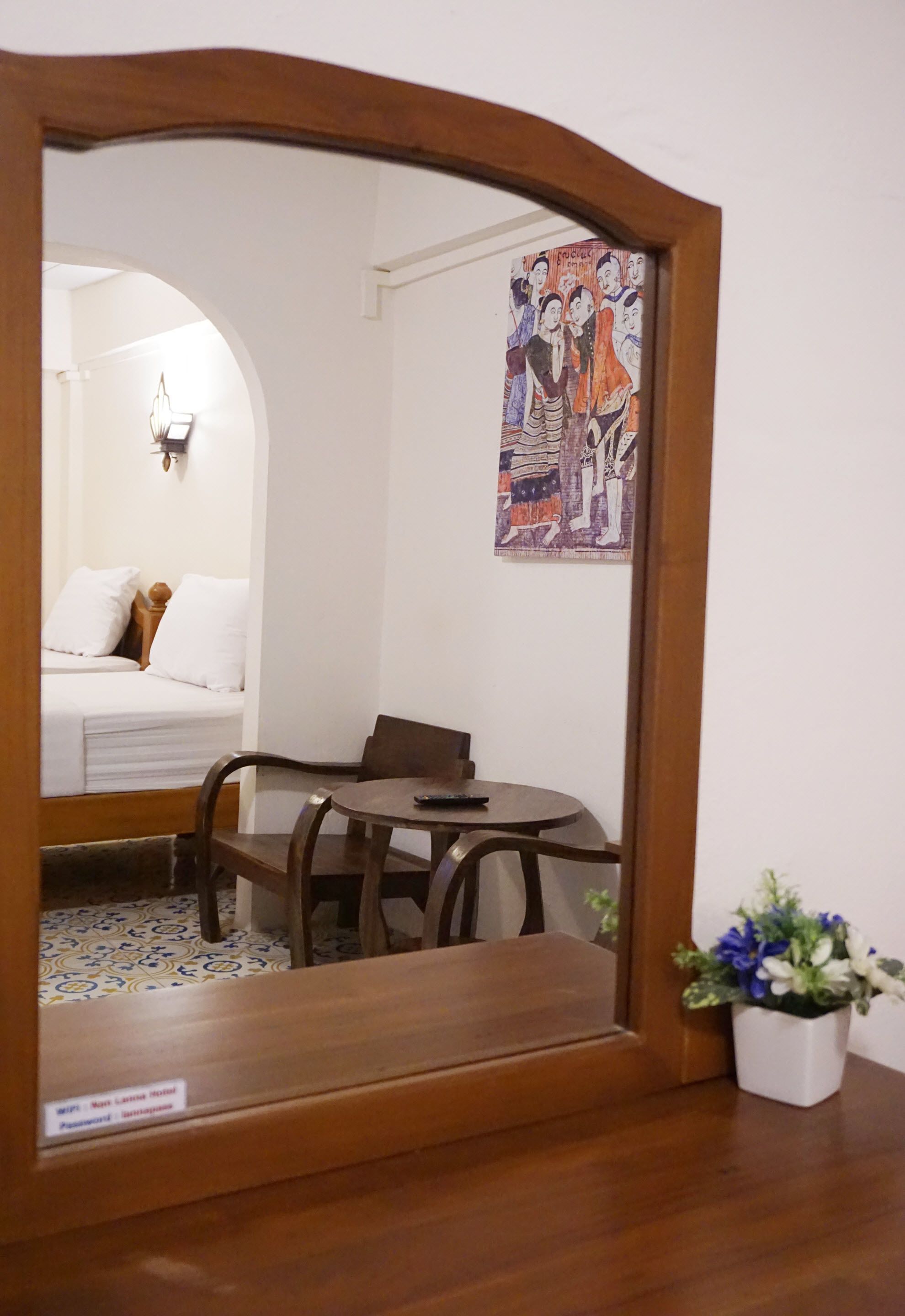 A room with a bed, a round table, and a chair. There is a painting on the wall and a potted plant on the floor.