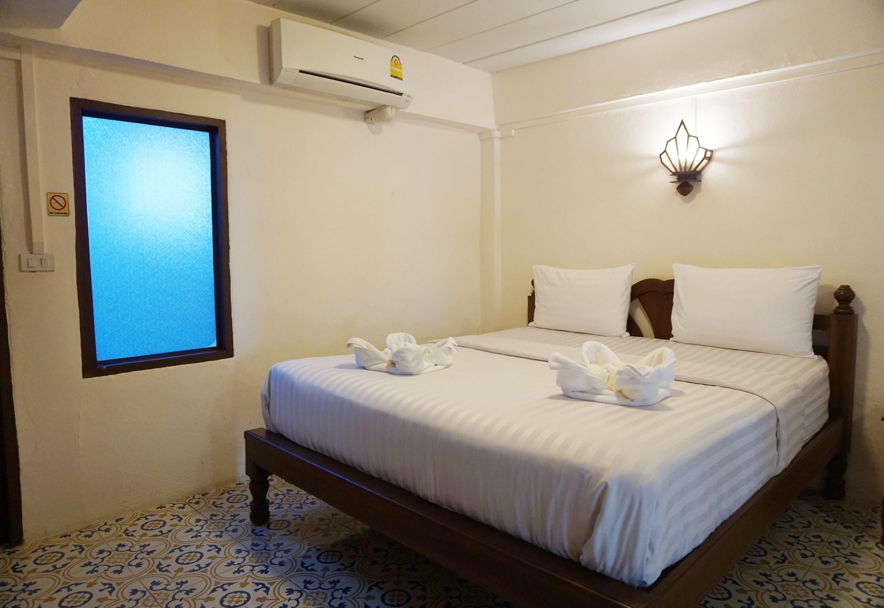 A hotel room with a bed, a window with blinds, an air conditioning unit, and a wall-mounted light fixture.