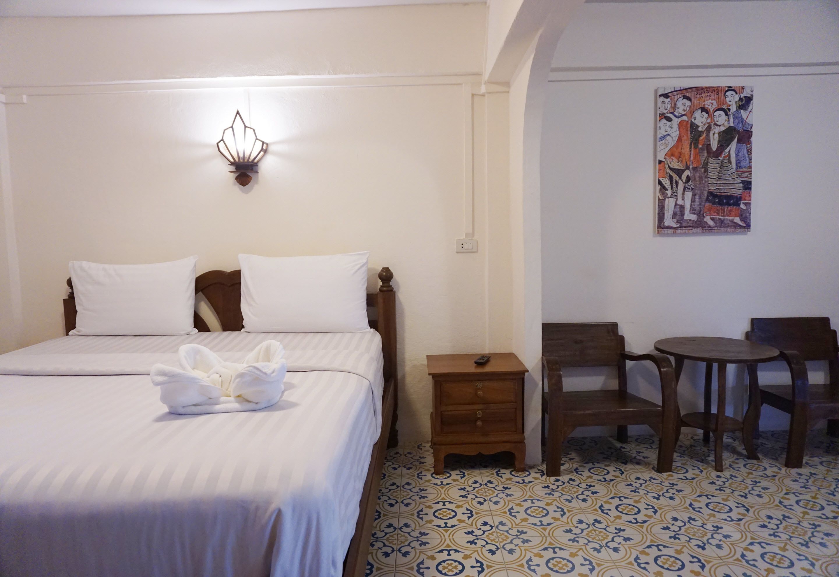 A hotel room with a bed, a nightstand, a chair, a small table, and a wall-mounted light fixture.