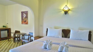 Quarto de hotel com cama bem arrumada, escrivaninha com cadeira, mesa pequena e luminária fixada na parede.