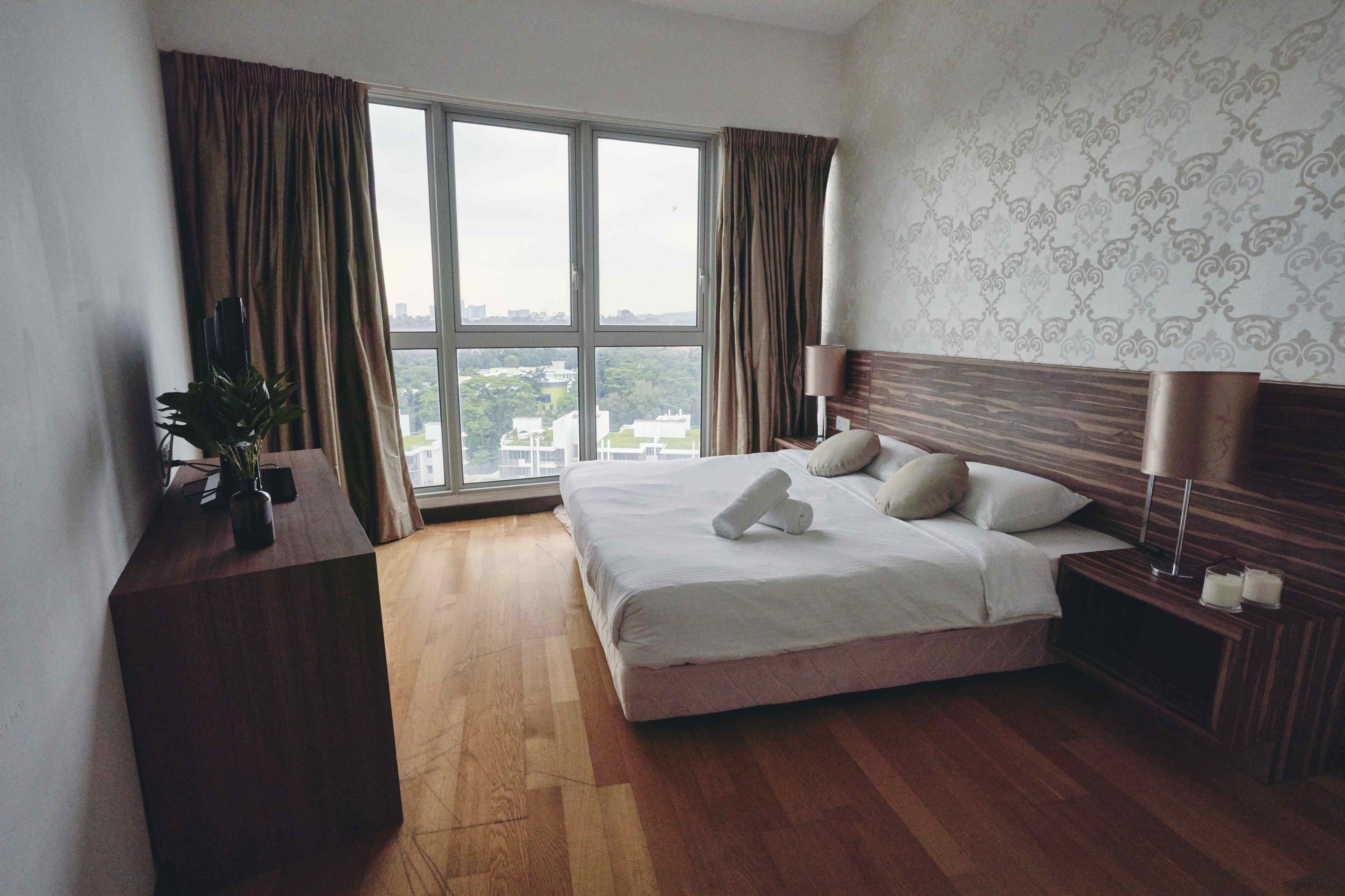 A hotel room with a large bed, a wooden desk, a potted plant, and a view of the city through large windows.