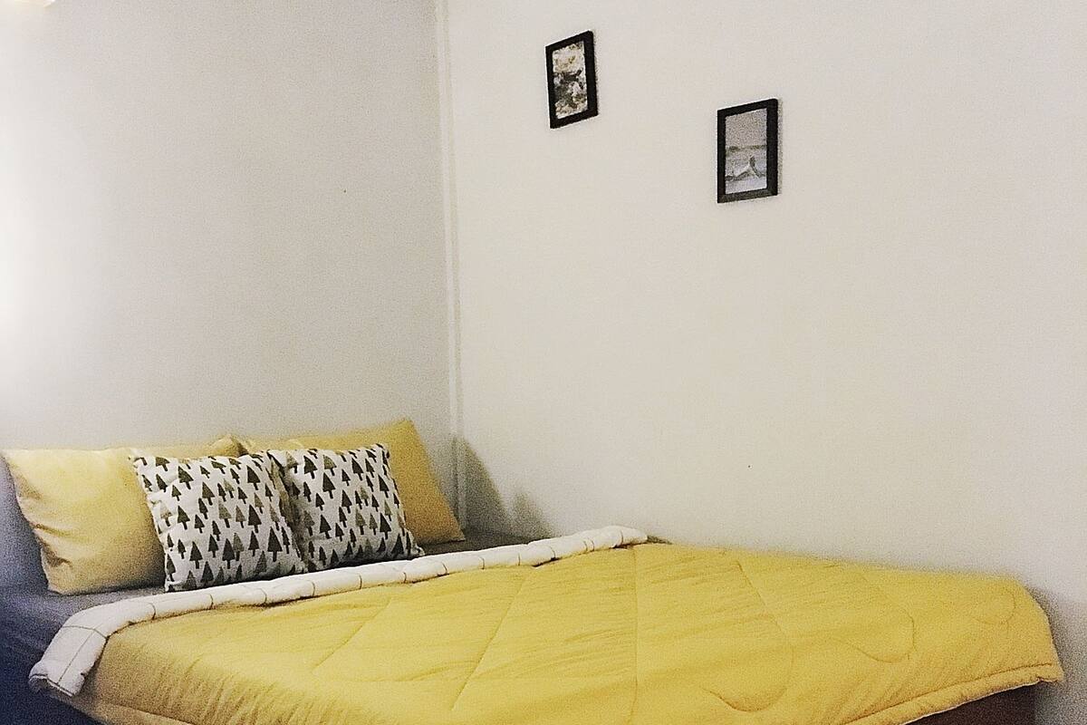 A bed with a yellow quilt, a black and white patterned pillow, and a black rug on a tiled floor.