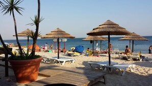 Een strand met rieten parasols, ligstoelen en een houten terras.
