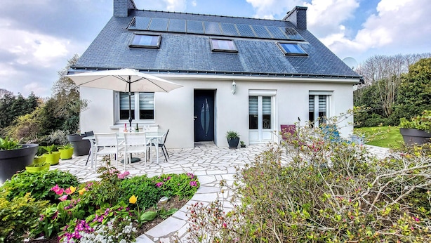Een tweelaags huis met een betegeld terras, een eethoek met tuinmeubels en een tuin vol planten en bloemen.