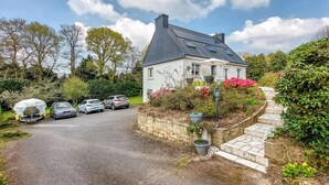 Een wit huis met een steil dak, geparkeerde auto's en een stenen muur.