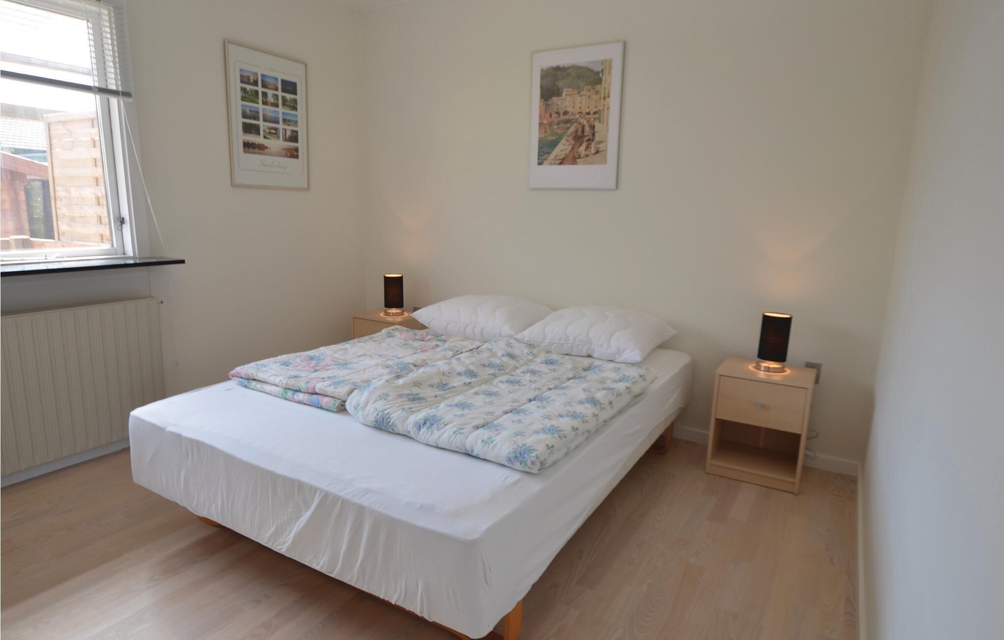 A bedroom with a bed, two bedside tables, a window, and two framed pictures on the wall.
