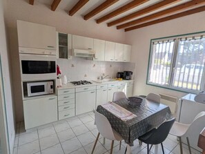 Une cuisine compacte avec des meubles blancs, une cuisinière à gaz, un four à micro-ondes et une machine à café. On y trouve une table à manger avec des chaises et une fenêtre avec des stores.