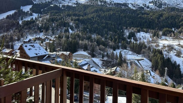 A mountainous landscape with snow-covered peaks and a forested valley. There are wooden buildings with sloped roofs, likely part of a resort or lodge.