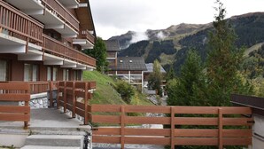 A multi-story building with wooden balconies, a stone staircase leading to the entrance, and a view of a mountainous landscape in the background.