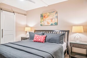 Un lit bien fait, recouvert d’une couverture grise, avec un coussin à motifs rouges et une tête de lit en bois. Deux tables de chevet avec des lampes se trouvent de chaque côté du lit, et un tableau représentant un paysage tropical est accroché au-dessus.