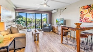 A modern living room with a sofa, a coffee table, a dining area with stools, a ceiling fan, and a large window offering a view of the outdoors. - GORGEOUS REMODEL-WELL EQUIPPED-MODERN LUXURY-AC THROUGHOUT (Kihei)