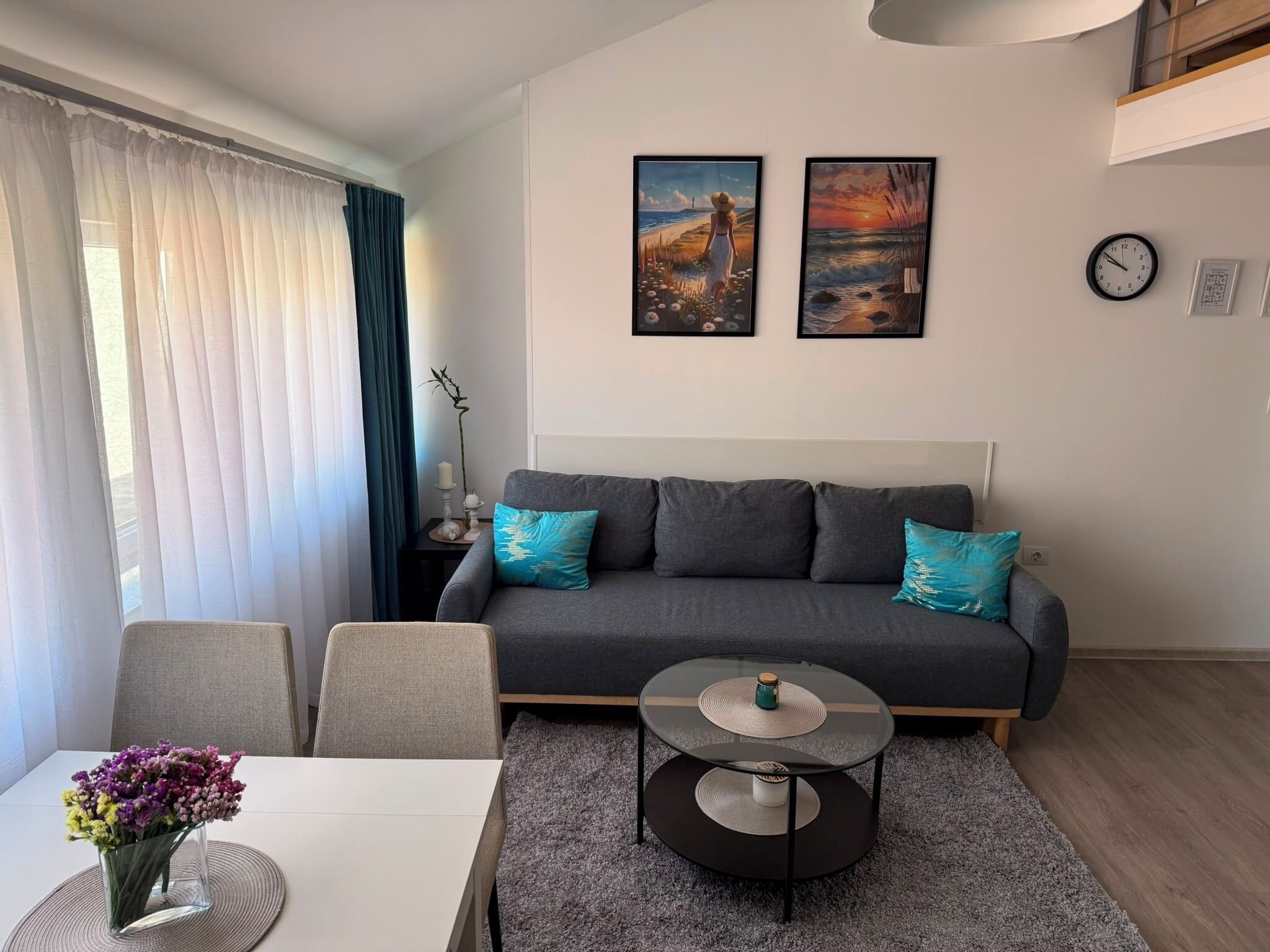 A modern living room with a grey sofa, two teal cushions, a round coffee table, and a dining area with a white table and a vase of flowers.