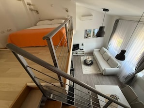 Un apartamento moderno con una escalera, una cama con una manta naranja, una sala de estar con sofá y televisor, y una mesa de comedor.