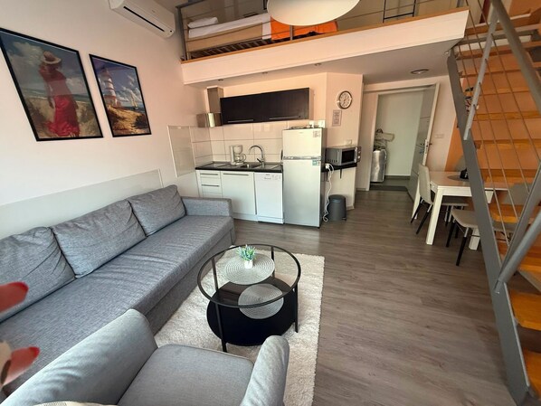 Un apartamento moderno con un sofá gris, una pequeña mesa redonda para café, una cocina y una mesa de comedor.