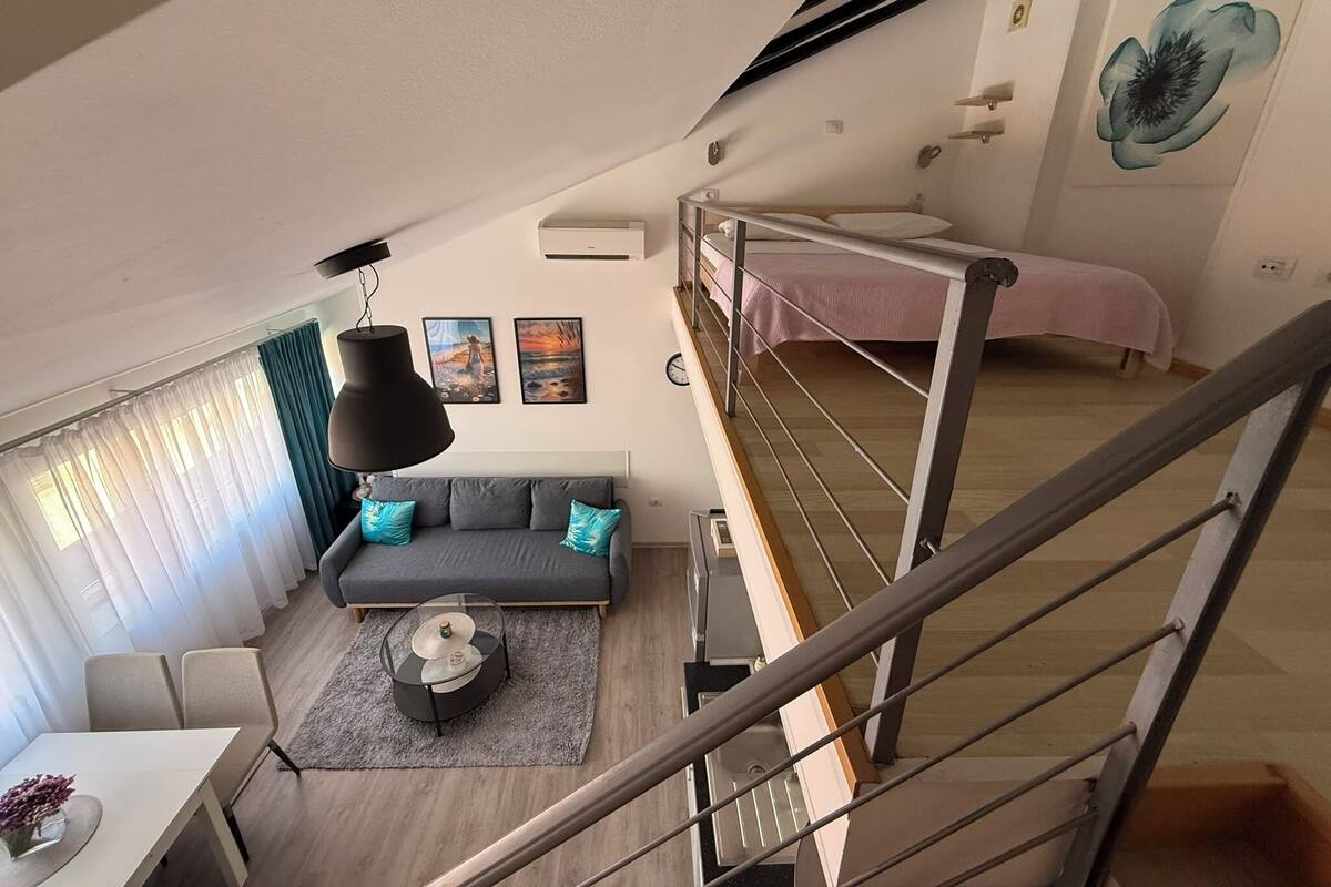 A modern apartment with a loft-style bedroom, a dining area, and a living room with a sofa and artwork.